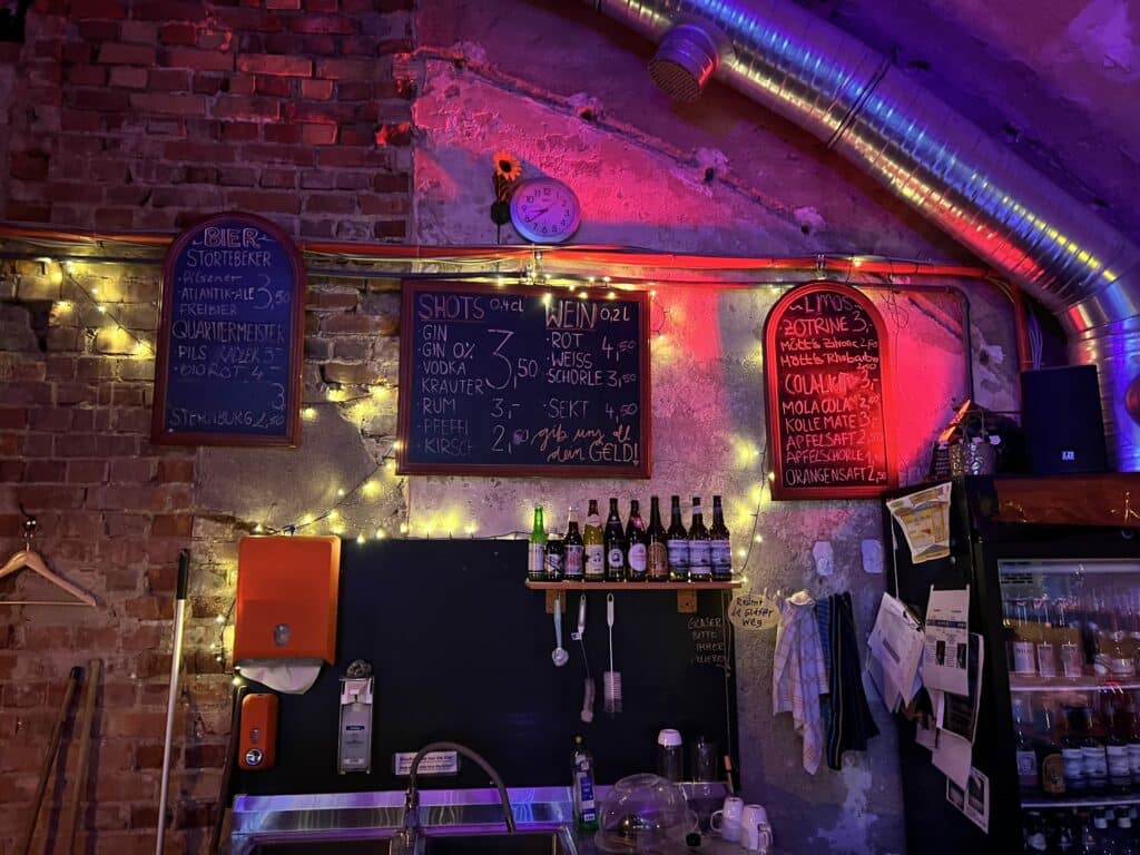 Eine kleine chaotische Bar. Handgeschriebene Tafeln an der Wand bewerben verschiedene Getränke. Bunte Lichterketten beleuchten die unverputzten Klinkerwände.