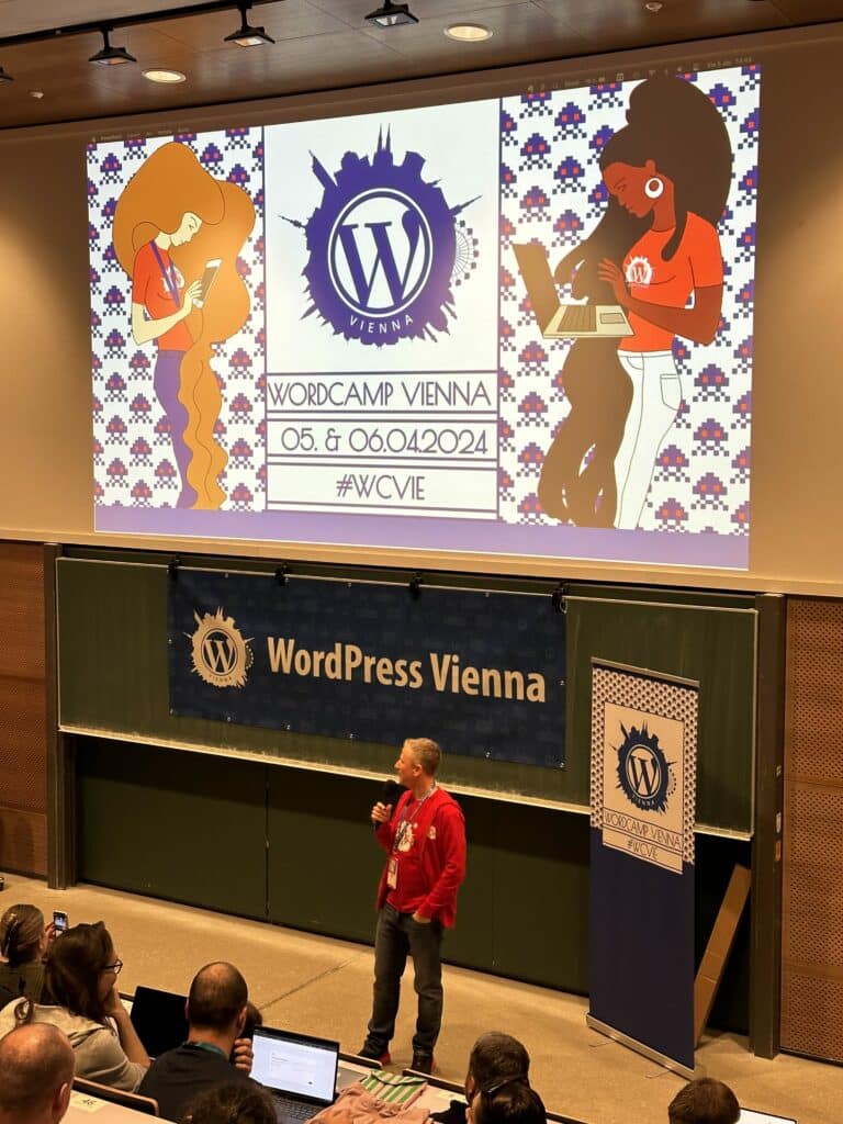 Foto einer Person in rotem Pullover unten in einem Hörsaar. Hinter der person ist der Schriftzug "WordPress Vienna" an der Wand